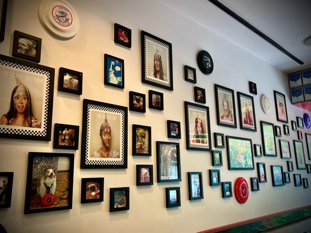 A wall of framed photos and frisbees, including portraits of people wearing ice cream cones on their heads, inside an Ashley’s Ice Cream shop