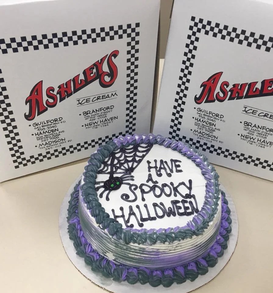 An Ashley’s ice cream cake decorated with a spider web and purple trim reading Have a Spooky Halloween, in front of checkered Ashley’s cake boxes