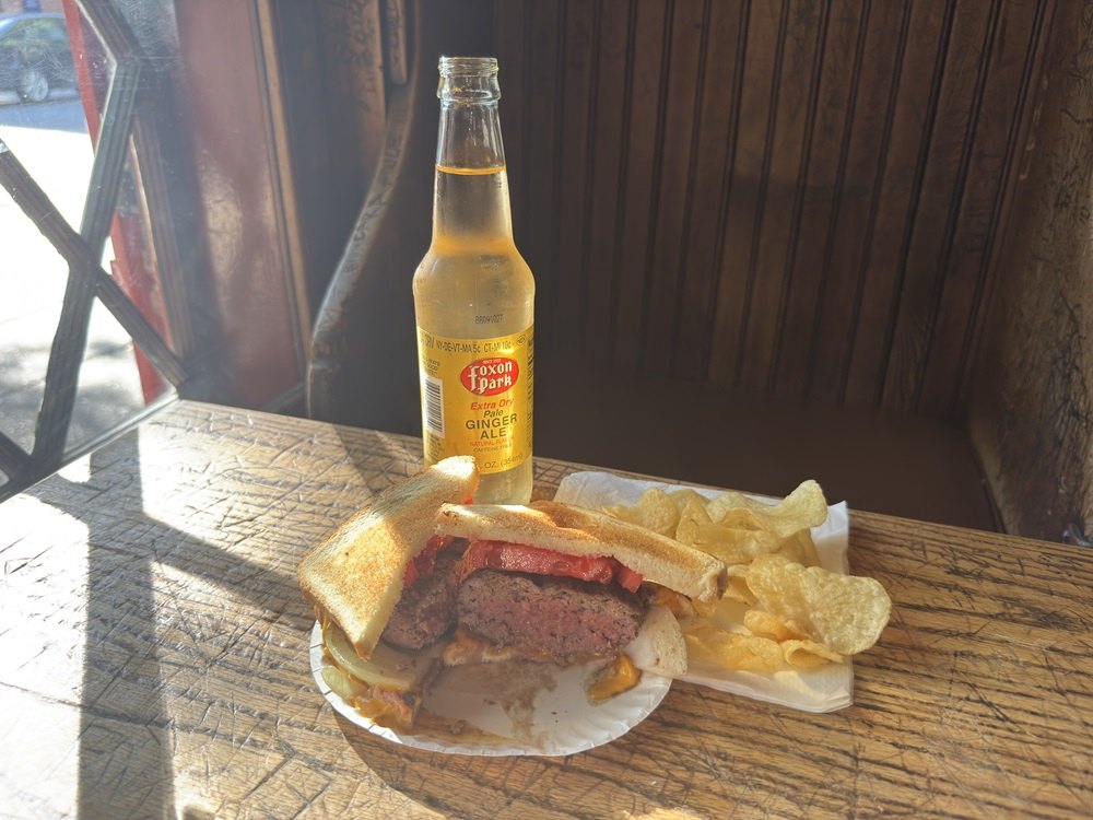 A Louis’ Lunch burger with chips and a soda on a wooden table