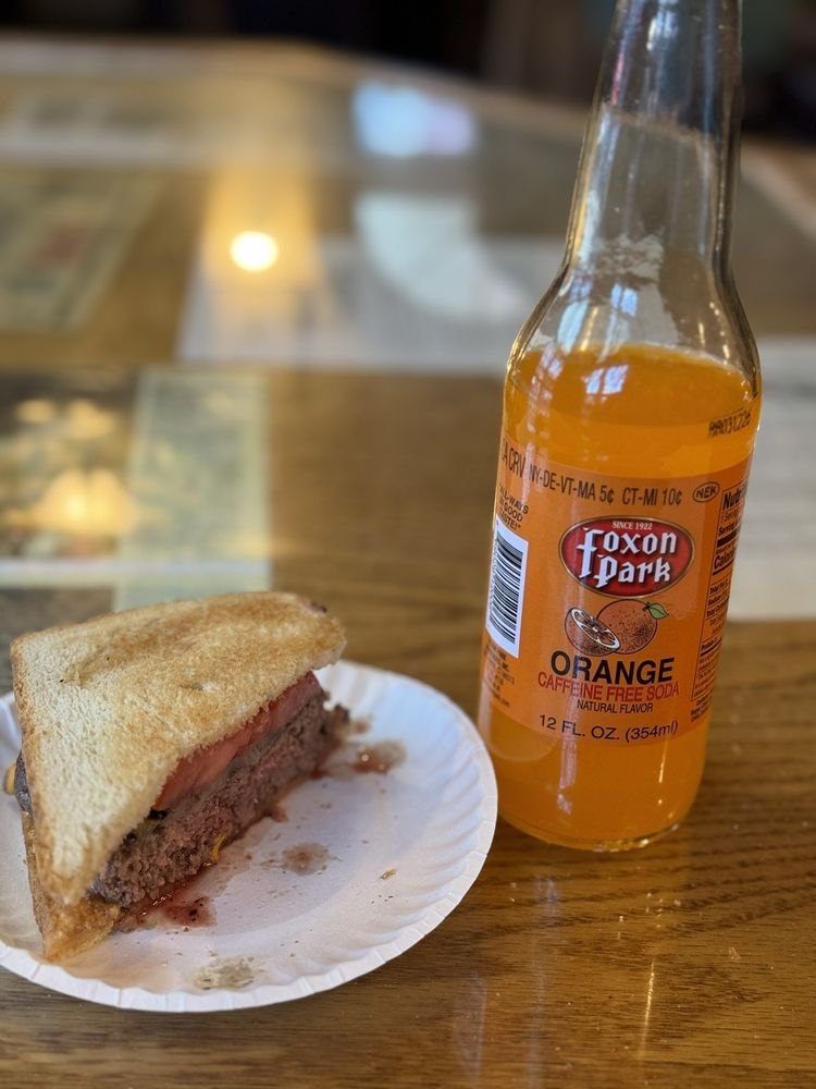 A Louis’ Lunch burger on a paper plate next to a bottle of Foxon Park orange soda