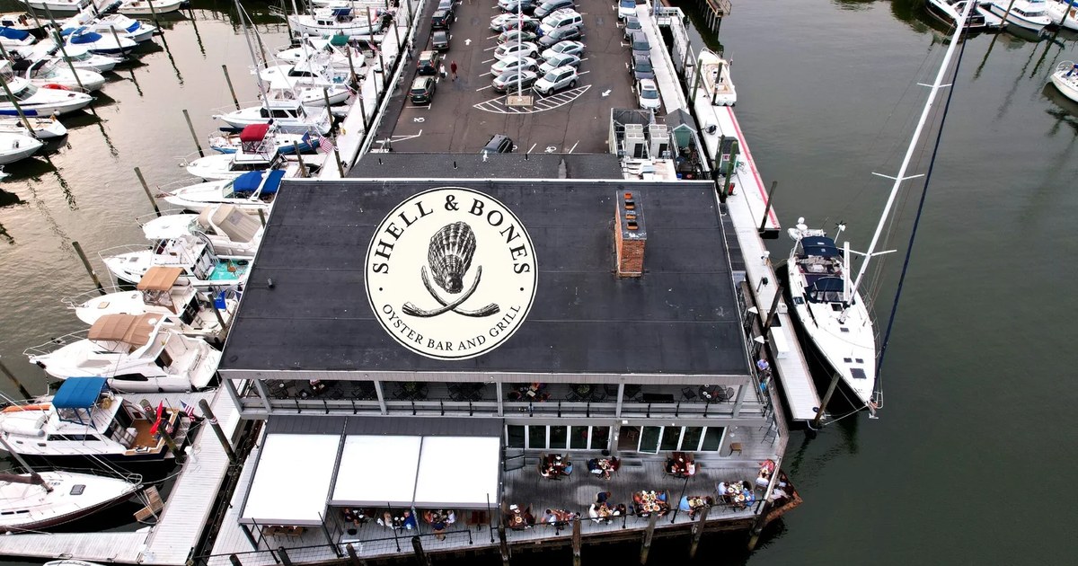Aerial view of Shell & Bones Oyster Bar and Grill on New Haven Harbor in City Point, with its logo painted on the roof and boats docked alongside