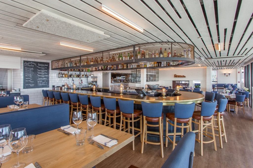 The long main bar at Shell & Bones with blue bar stools and ship-style lights