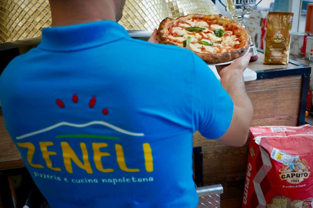 A Zeneli pizzaiolo in a blue shirt holding a Margherita in front of the gold wood-fired oven